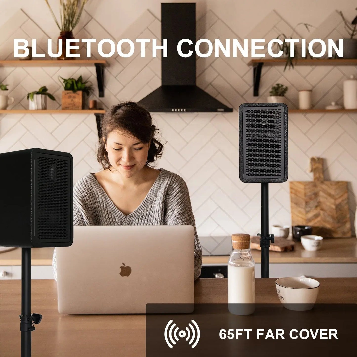 A woman sits at a kitchen table with a laptop, smiling at the screen, while using two tall, black, rectangular speakers on stands; text above reads 'BLUETOOTH CONNECTION' and at the bottom right, an icon indicates '65FT FAR COVER' range.