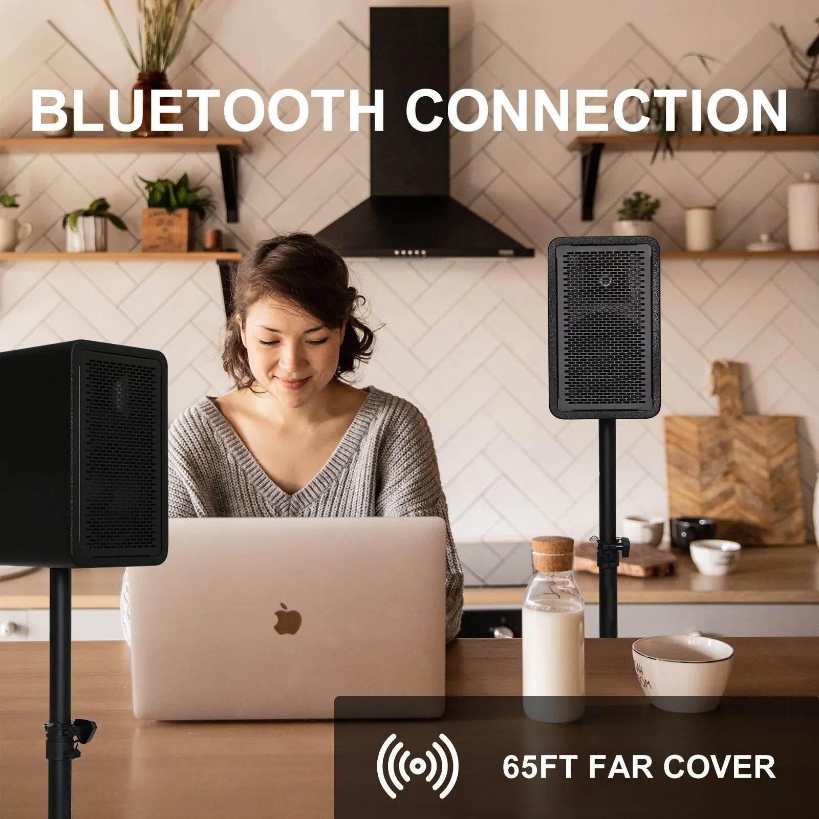 A woman sits at a kitchen table with a laptop, smiling at the screen, while using two tall, black, rectangular speakers on stands; text above reads 'BLUETOOTH CONNECTION' and at the bottom right, an icon indicates '65FT FAR COVER' range.